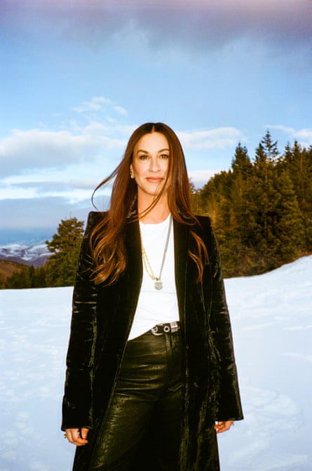 Alanis Morissette against a snowy mountain scene wearing a long black coat, white T-shirt and dark trousers