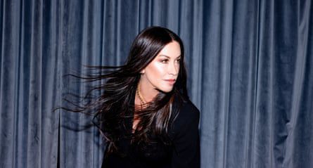 Portrait of Alanis Morissette in a dark jacket against a dark curtain with her shiny dark hair billowing about her