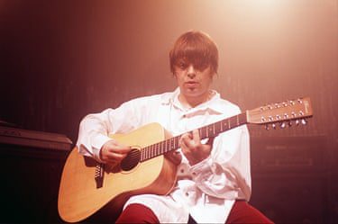 Mani plays a 12-string acoustic guitar, sitting wearing a white shirt and red trousers.