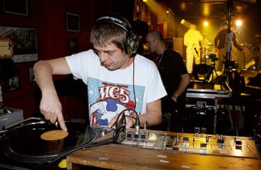 Mani places his finger on a vinyl record while standing in a DJ booth.